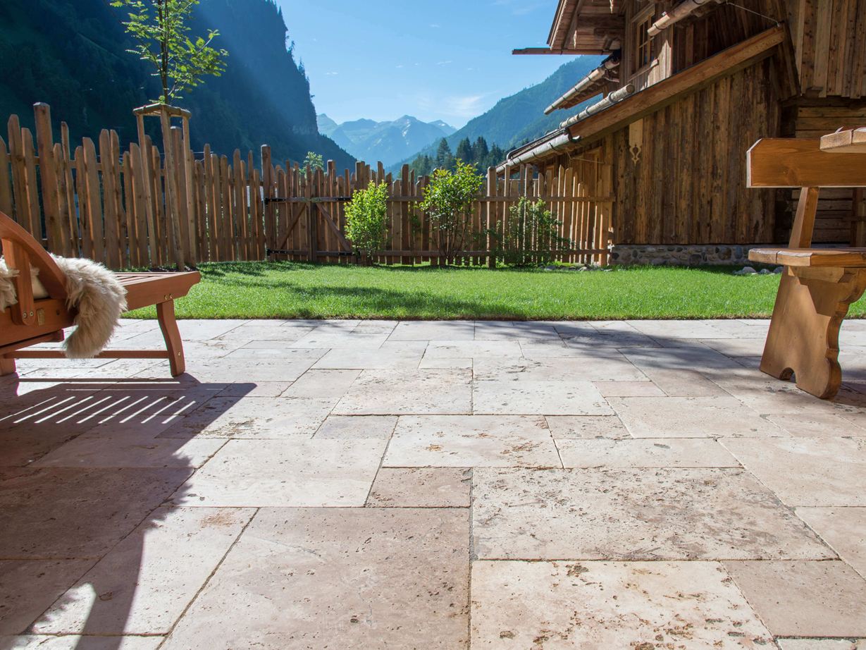 Des tons beiges doux et chauds à la lumière du soleil Terrasse en travertin Rustic avec meubles rustiques en bois, en arrière-plan une pelouse et une vue sur les montagnes