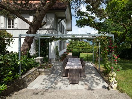 Terrasse Kandla Grey en grès avec un salon de jardin en bois, une fontaine fleurie et de la verdure tout autour