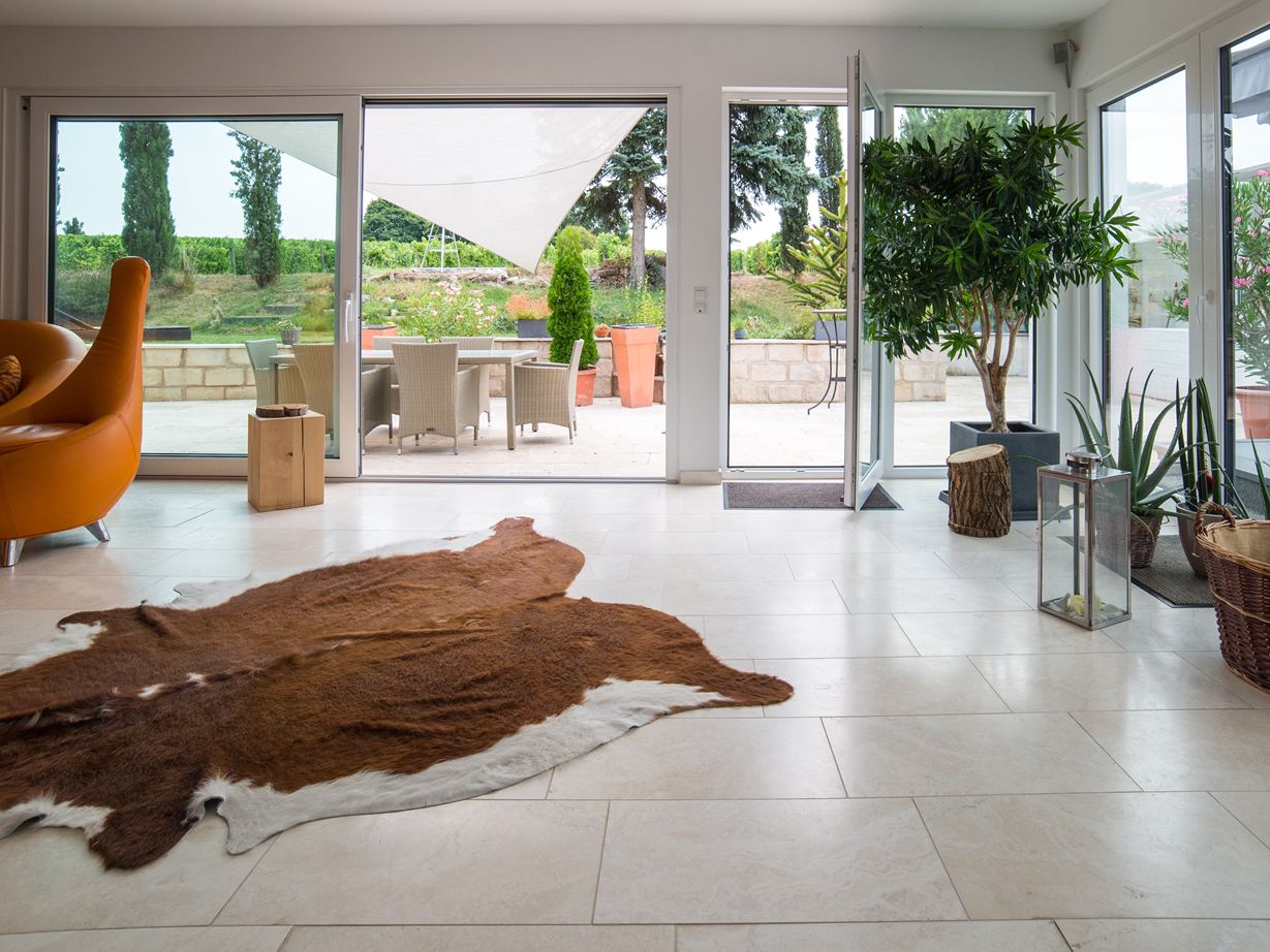 Ce travertin donne une note chic au mobilier. Baie vitrée séjour tapis en peau de bête plante fauteil orange carrelage Vanilla Rustica travertin